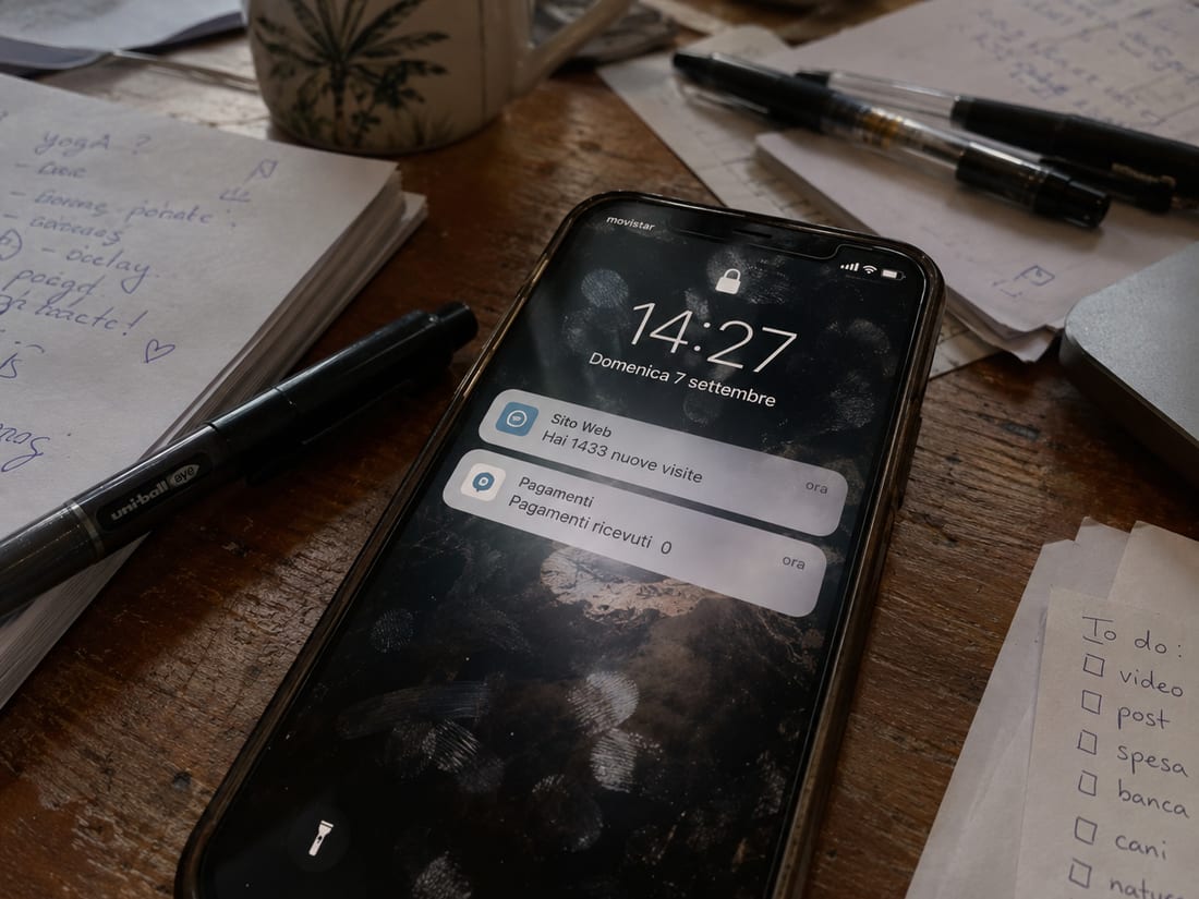 Un telefono sulla scrivania fra appunti e una lista di cose da fare: sullo schermo due notifiche, «Sito Web — hai 1433 nuove visite» e sotto «Pagamenti — pagamenti ricevuti 0»
